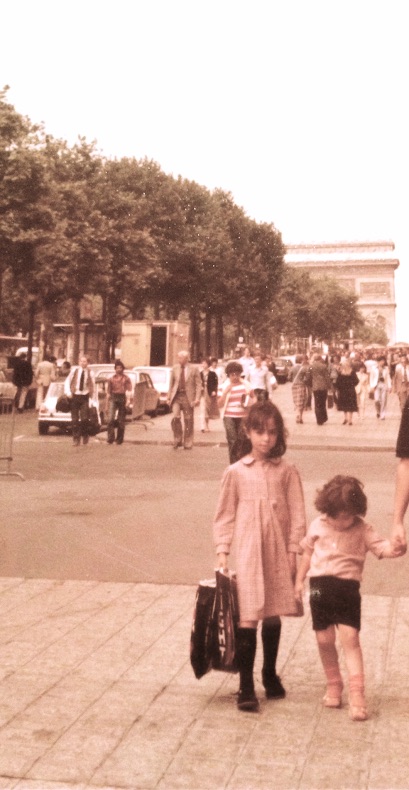 Two small girls walking hand in hand on a Paris sidewalk, the Arc de Triomphe behind them