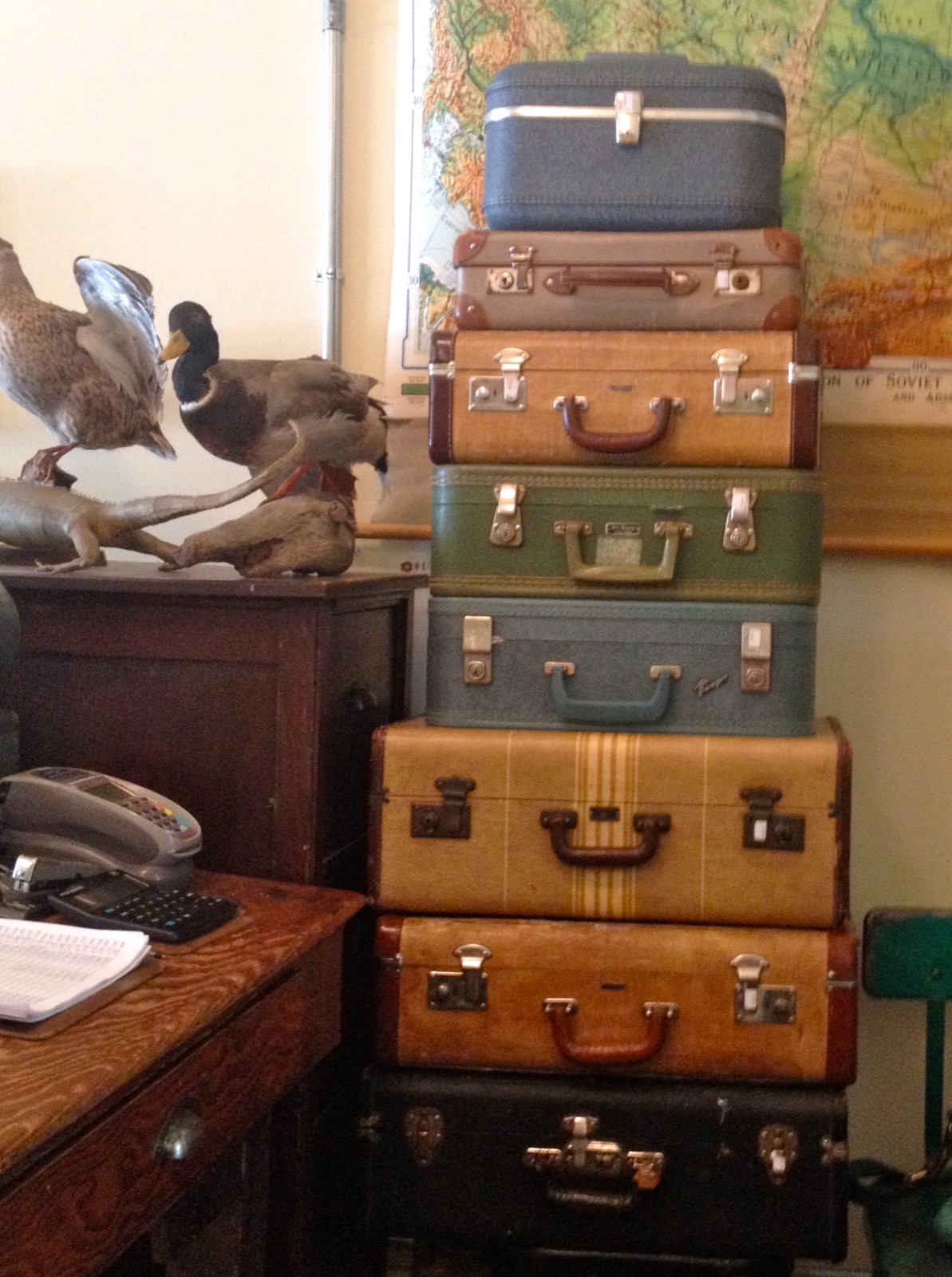 A tall stack of vintage suitcases beside an old desk, with ducks and a wall map behind